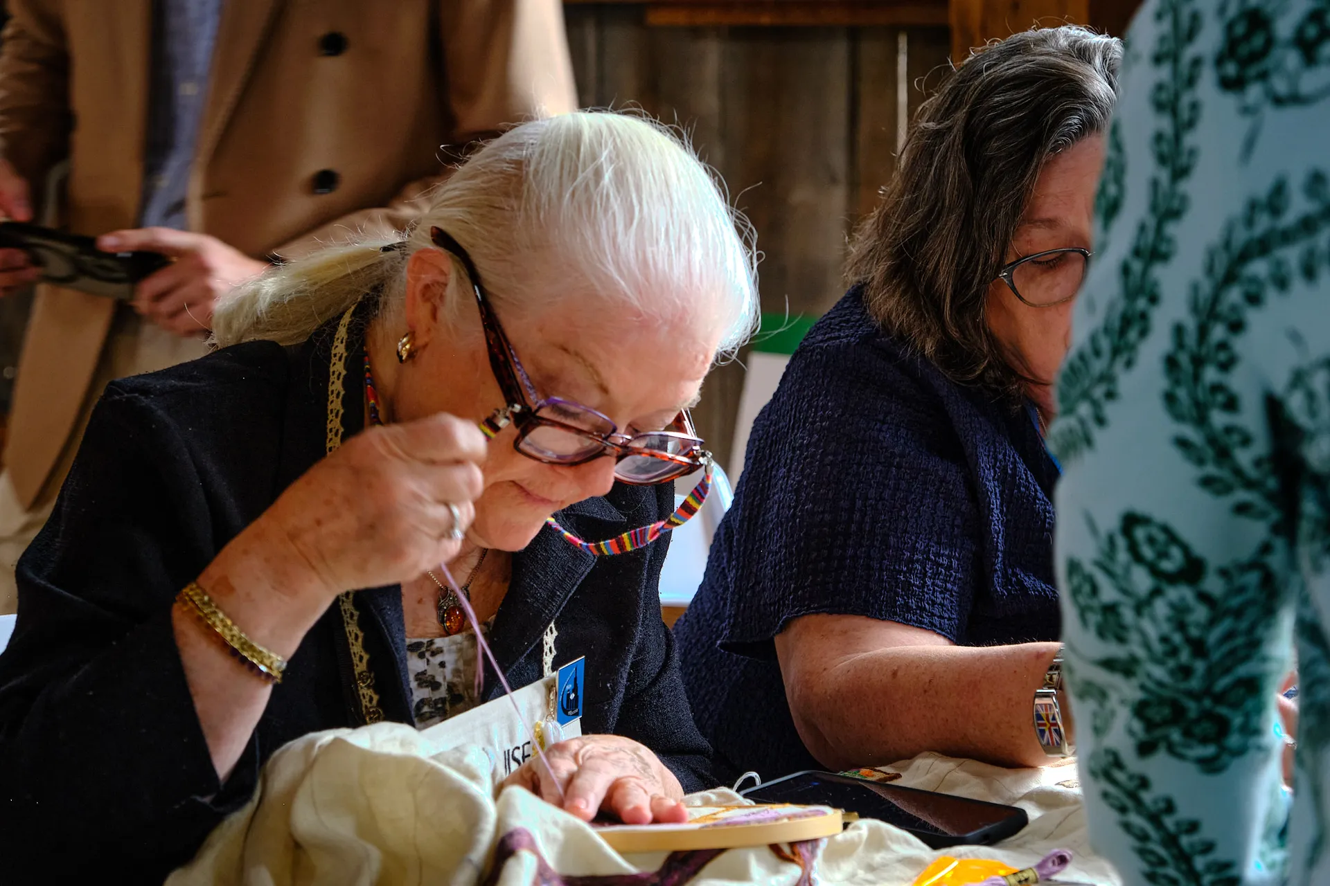 Two stitchers concentrate on their needlework, one drawing thread through the linen